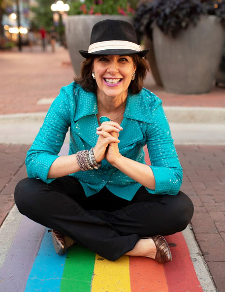 Tama Kieves sitting on a rainbow colored sidewalk smiling at the camera